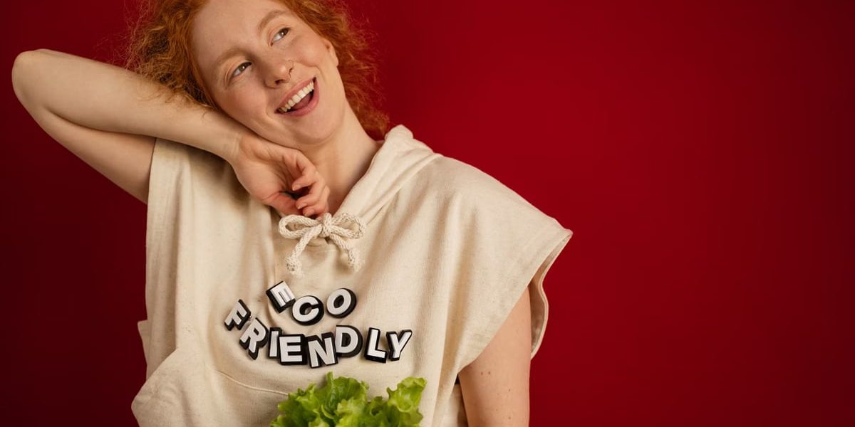 Cheerful woman with red hair in eco-friendly attire holding fresh lettuce against a vibrant red background.