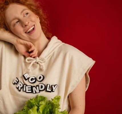Cheerful woman with red hair in eco-friendly attire holding fresh lettuce against a vibrant red background.